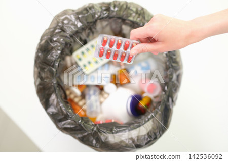 Hand discarding blister pack of red capsules into trash bin filled with assorted medicines, showing medicine disposal, drug waste, expired pills, household cleanup, safety, and healthcare waste Hand discarding blister pack of red capsules into trash bin filled with assorted medicines, showing medicine disposal, drug waste, expired pills, household cleanup, safety, and healthcare waste 142536092