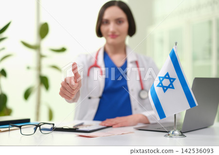 Smiling female doctor in white coat giving thumbs up beside Israeli flag on desk in bright clinic office, with stethoscope, laptop, glasses, and papers in soft daylight Smiling female doctor in white coat giving thumbs up beside Israeli flag on desk in bright clinic office, with stethoscope, laptop, glasses, and papers in soft daylight 142536093