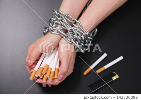 Bound hands holding cigarettes with metal chain on dark background, symbolizing smoking addiction, quitting struggle, nicotine dependence, unhealthy habit, restraint, recovery, control, and anti 142536099