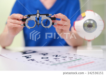Person holding optometry trial frame during eye examination with vision chart on desk and eye model nearby, healthcare setting focused on eyesight testing, lens fitting, and visual assessment Person holding optometry trial frame during eye examination with vision chart on desk and eye model nearby, healthcare setting focused on eyesight testing, lens fitting, and visual assessment 142536151