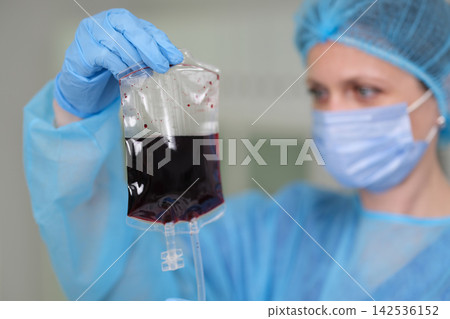 Masked healthcare worker in blue protective gown and gloves holding blood bag with dark fluid for transfusion inspection in clinical environment, with healthcare, medical, nurse, doctor detail 142536152