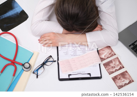 Stressed doctor covering face over medical paperwork, clipboard, eyeglasses, stethoscope, and ultrasound printouts on white desk, viewed from above in clinical office setting Stressed doctor covering face over medical paperwork, clipboard, eyeglasses, stethoscope, and ultrasound printouts on white desk, viewed from above in clinical office setting 142536154