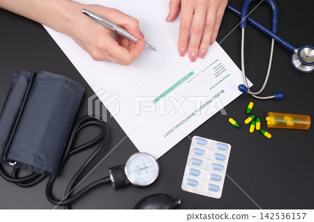 Hands writing blood pressure treatment notes on medical form beside stethoscope, cuff, pills, blister pack and gauge on dark desk, showing healthcare planning, medication management and home Hands writing blood pressure treatment notes on medical form beside stethoscope, cuff, pills, blister pack and gauge on dark desk, showing healthcare planning, medication management and home 142536157