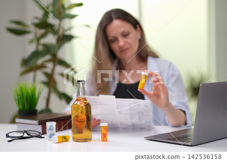 Woman reviewing prescription medication at desk, holding pill bottle beside laptop, reading leaflet with glasses, bottles and medicine containers in home workspace, healthcare decision and self care Woman reviewing prescription medication at desk, holding pill bottle beside laptop, reading leaflet with glasses, bottles and medicine containers in home workspace, healthcare decision and self care 142536158