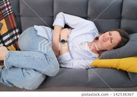 Woman lies on sofa holding abdomen with hands suffering from pain Woman lies on sofa holding abdomen with hands suffering from pain 142536367