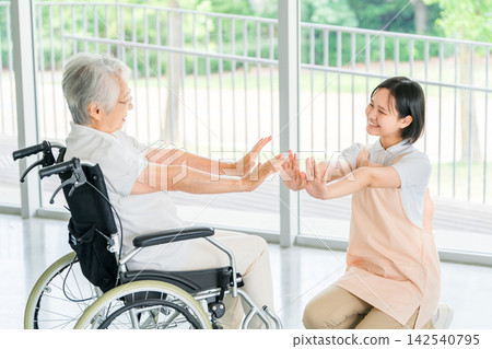 功能性訓練:一位坐在輪椅上的老年婦女和她的護理人員面帶微笑地進行鍛鍊。 功能性訓練:一位坐在輪椅上的老年婦女和她的護理人員面帶微笑地進行鍛鍊。 142540795