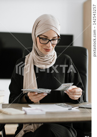A professional woman wearing a hijab and glasses sits at a desk, meticulously counting US dollar bills, symbolizing financial management and business A professional woman wearing a hijab and glasses sits at a desk, meticulously counting US dollar bills, symbolizing financial management and business 142543257