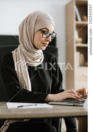 A professional Muslim woman wearing a hijab and glasses is focused on her work while typing on a laptop at her desk A professional Muslim woman wearing a hijab and glasses is focused on her work while typing on a laptop at her desk 142543357