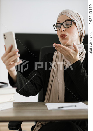 A woman wearing a hijab and glasses is on a video call, blowing a kiss to the phone screen while sitting at her desk A woman wearing a hijab and glasses is on a video call, blowing a kiss to the phone screen while sitting at her desk 142543359