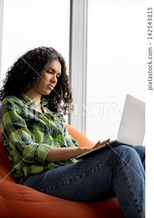 A young woman with curly hair sits comfortably in an orange beanbag chair, focused on her laptop screen A young woman with curly hair sits comfortably in an orange beanbag chair, focused on her laptop screen 142543815