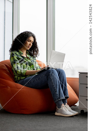 A young woman with curly hair sits comfortably on a large orange bean bag chair, focused on her laptop in a bright, modern space 142543824