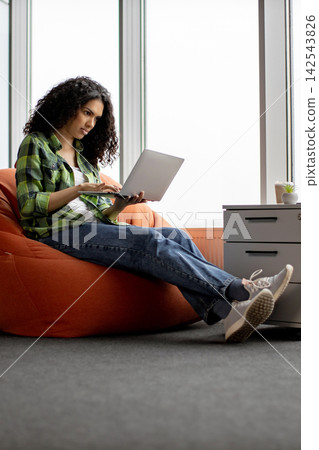 A young woman with curly hair sits comfortably on an orange beanbag chair, focused on her laptop A young woman with curly hair sits comfortably on an orange beanbag chair, focused on her laptop 142543826