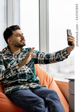 A young man with a beard wearing a plaid shirt sits in an orange beanbag chair and records a video on his smartphone A young man with a beard wearing a plaid shirt sits in an orange beanbag chair and records a video on his smartphone 142543845