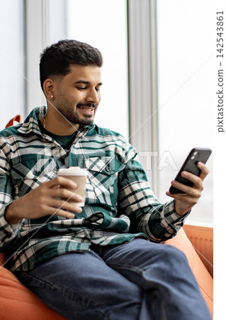 A young man in a plaid shirt holds a coffee cup and looks at his smartphone while sitting comfortably A young man in a plaid shirt holds a coffee cup and looks at his smartphone while sitting comfortably 142543861