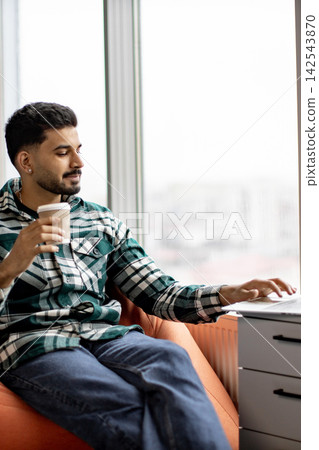 A young man with a beard and a plaid shirt sits comfortably in an armchair, holding a coffee cup while typing on his laptop near a large window A young man with a beard and a plaid shirt sits comfortably in an armchair, holding a coffee cup while typing on his laptop near a large window 142543870