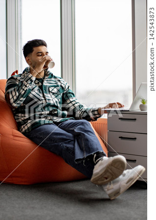 A young man in casual attire enjoys a beverage while working on his laptop from a comfortable beanbag chair near a large window A young man in casual attire enjoys a beverage while working on his laptop from a comfortable beanbag chair near a large window 142543873