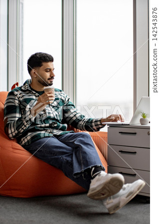 A young man in a plaid shirt relaxes in an orange beanbag chair while working on his laptop and holding a coffee cup 142543876