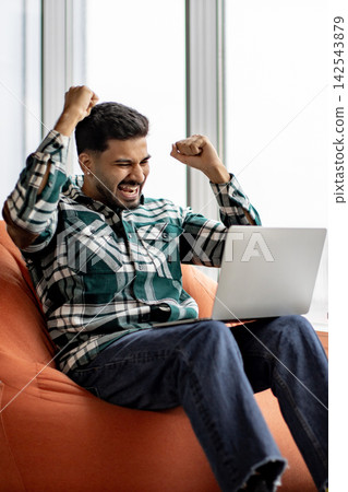 A young man with a beard and dark hair, wearing a plaid shirt and jeans, sits on an orange beanbag chair and celebrates enthusiastically while looking at his laptop screen A young man with a beard and dark hair, wearing a plaid shirt and jeans, sits on an orange beanbag chair and celebrates enthusiastically while looking at his laptop screen 142543879