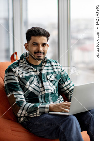 A young man with a beard and a plaid shirt sits comfortably in an orange chair, typing on a laptop near a window A young man with a beard and a plaid shirt sits comfortably in an orange chair, typing on a laptop near a window 142543905