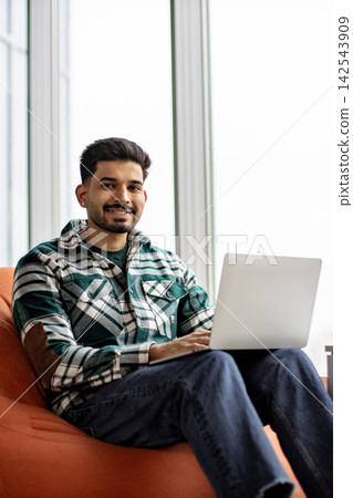 A smiling young man with a beard and dark hair sits comfortably on an orange beanbag chair while typing on a silver laptop A smiling young man with a beard and dark hair sits comfortably on an orange beanbag chair while typing on a silver laptop 142543909