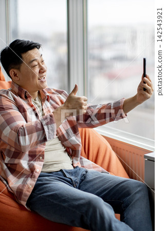 A cheerful Asian man sits comfortably in a beanbag chair, holding a smartphone to take a selfie while giving a thumbs-up gesture 142543921