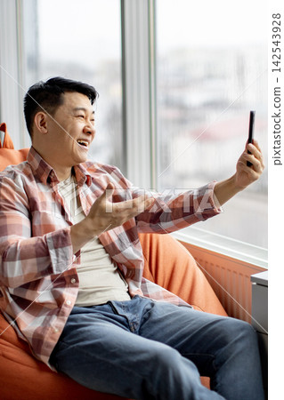 A cheerful Asian man in a plaid shirt is holding a smartphone and video calling while sitting comfortably in an armchair by the window A cheerful Asian man in a plaid shirt is holding a smartphone and video calling while sitting comfortably in an armchair by the window 142543928