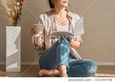 A woman enjoying a book while sipping coffee in a warm, sunlit room. A woman enjoying a book while sipping coffee in a warm, sunlit room. 142544796