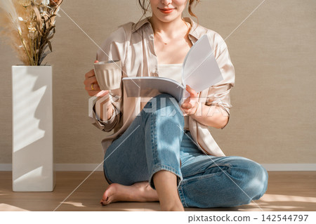 A woman enjoying a book while sipping coffee in a warm, sunlit room. A woman enjoying a book while sipping coffee in a warm, sunlit room. 142544797