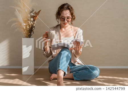 A woman enjoying a book while sipping coffee in a warm, sunlit room. A woman enjoying a book while sipping coffee in a warm, sunlit room. 142544799