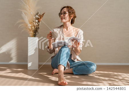 A woman enjoying reading and coffee barefoot in a warm, sunlit room. A woman enjoying reading and coffee barefoot in a warm, sunlit room. 142544801