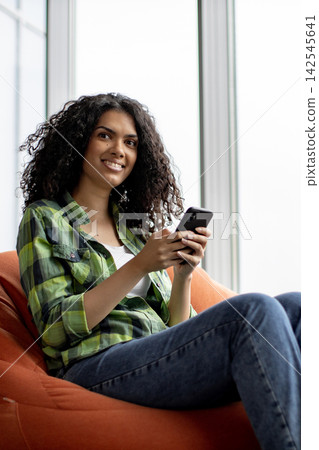 A young woman with curly hair smiles while holding a smartphone and sitting on an orange beanbag chair near a window A young woman with curly hair smiles while holding a smartphone and sitting on an orange beanbag chair near a window 142545641