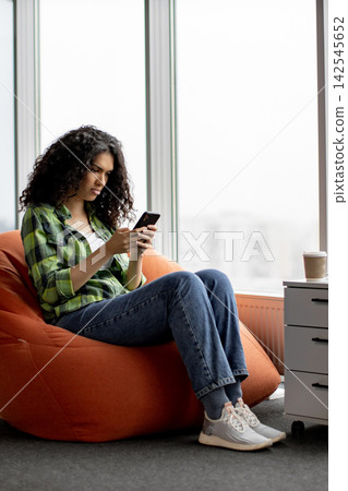 A young woman with curly hair sits comfortably in an orange beanbag chair, engrossed in her mobile phone 142545652