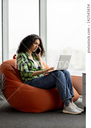 A young woman with curly hair sits on a beanbag chair, talking on her phone while typing on a laptop, with large windows in the background A young woman with curly hair sits on a beanbag chair, talking on her phone while typing on a laptop, with large windows in the background 142545654