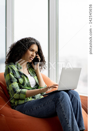 A young woman with curly hair sits comfortably in an orange beanbag chair, engaged in a phone conversation while typing on her laptop 142545656