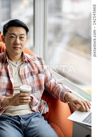 A man sits comfortably in an armchair by the window, holding a coffee cup and working on his laptop A man sits comfortably in an armchair by the window, holding a coffee cup and working on his laptop 142545668