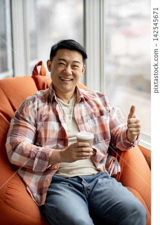 A cheerful Asian man in a plaid shirt gives a thumbs up while holding a coffee cup and relaxing in a beanbag chair by the window A cheerful Asian man in a plaid shirt gives a thumbs up while holding a coffee cup and relaxing in a beanbag chair by the window 142545671