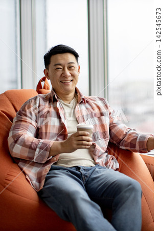 A smiling Asian man in a plaid shirt sits comfortably in an orange beanbag chair holding a disposable coffee cup A smiling Asian man in a plaid shirt sits comfortably in an orange beanbag chair holding a disposable coffee cup 142545673