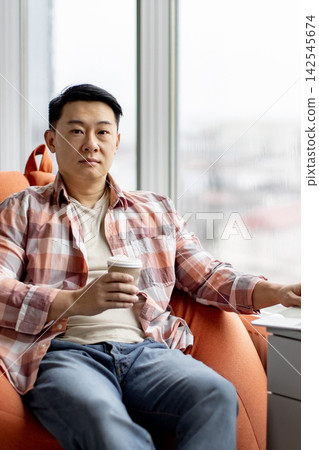 A man in a plaid shirt and jeans sits in an orange chair holding a disposable coffee cup while looking towards the camera with a neutral expression A man in a plaid shirt and jeans sits in an orange chair holding a disposable coffee cup while looking towards the camera with a neutral expression 142545674