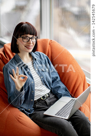 A smiling woman wearing glasses and a denim shirt sits comfortably in an orange beanbag chair, holding a laptop and giving an okay sign 142545679