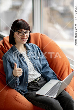 A young woman with glasses and a denim shirt gives a thumbs-up while working on a laptop, seated comfortably in a beanbag chair by a window A young woman with glasses and a denim shirt gives a thumbs-up while working on a laptop, seated comfortably in a beanbag chair by a window 142545680