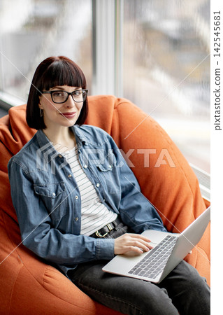 A young woman with glasses and a denim shirt sits comfortably in a large orange beanbag chair, typing on her laptop near a window 142545681
