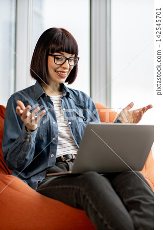 A young woman with glasses and a bob haircut smiles while gesturing during a video call on her laptop, seated comfortably in an orange beanbag chair A young woman with glasses and a bob haircut smiles while gesturing during a video call on her laptop, seated comfortably in an orange beanbag chair 142545701