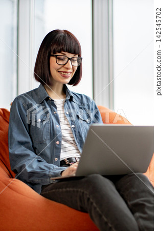A young woman with glasses and a bob haircut smiles while working on her laptop computer, seated comfortably on an orange beanbag chair A young woman with glasses and a bob haircut smiles while working on her laptop computer, seated comfortably on an orange beanbag chair 142545702
