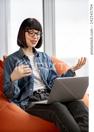 A young woman with a bob haircut and glasses is actively engaged in a video call on her laptop while sitting comfortably in an orange beanbag chair A young woman with a bob haircut and glasses is actively engaged in a video call on her laptop while sitting comfortably in an orange beanbag chair 142545704
