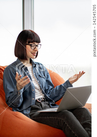 A young woman with glasses and a bob haircut is sitting comfortably on an orange beanbag chair while working on her laptop A young woman with glasses and a bob haircut is sitting comfortably on an orange beanbag chair while working on her laptop 142545705