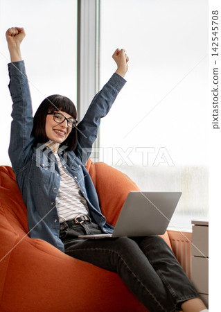 A young woman with glasses and a denim shirt raises her arms in triumph while sitting in an orange beanbag chair with a laptop A young woman with glasses and a denim shirt raises her arms in triumph while sitting in an orange beanbag chair with a laptop 142545708