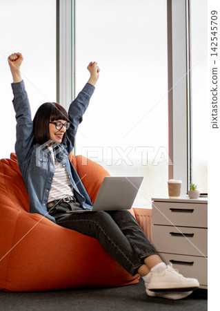 A young woman with glasses and a denim shirt sits on an orange beanbag chair, raising her arms in triumph while working on her laptop A young woman with glasses and a denim shirt sits on an orange beanbag chair, raising her arms in triumph while working on her laptop 142545709