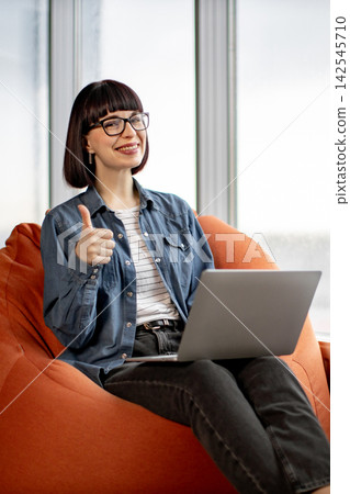 A young woman with glasses and a bob haircut sits comfortably on an orange beanbag chair, giving a thumbs-up gesture while using her laptop 142545710
