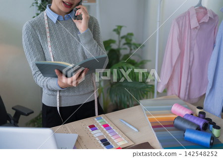 Asian fashion designer consulting client by phone while reviewing color swatches and fabric samples in home studio workspace Asian fashion designer consulting client by phone while reviewing color swatches and fabric samples in home studio workspace 142548252