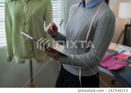 Asian fashion designer with tape measure reviewing digital tablet while examining green garment in studio workspace Asian fashion designer with tape measure reviewing digital tablet while examining green garment in studio workspace 142548253